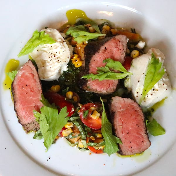 Seared beef slices with fresh mozzarella, corn, cherry tomatoes, and green herbs on a white plate.