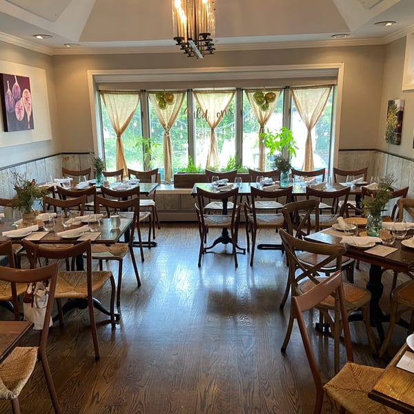 A dining room with wooden tables and chairs, set with white napkins and glasses, bathed in natural light.