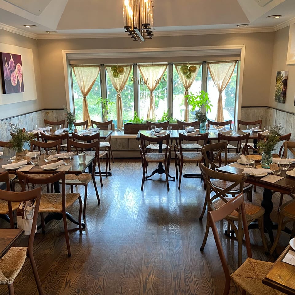 A dining room with wooden tables and chairs, set with white napkins and glasses, bathed in natural light.