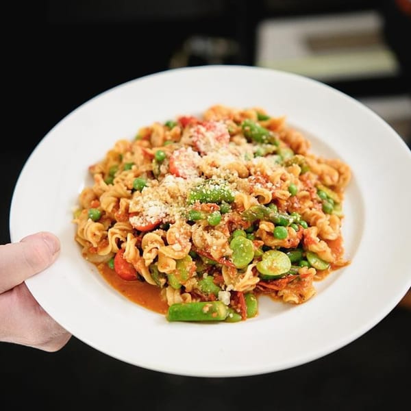 A hand holds a white plate of fusilli pasta with tomato sauce, green peas, and broad beans.