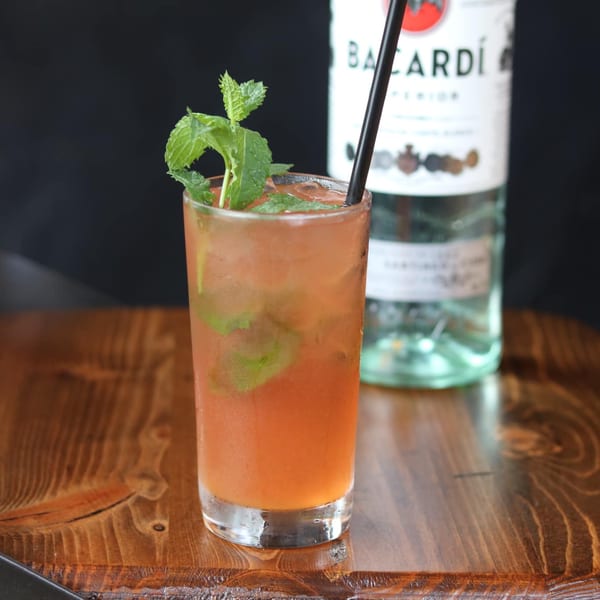 A tall glass of iced orange cocktail with mint and a black straw, with a Bacardi bottle behind it.