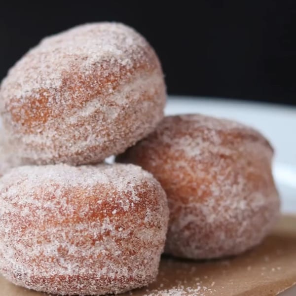 Three sugar-dusted donut holes are stacked on a white plate with a brown paper liner.