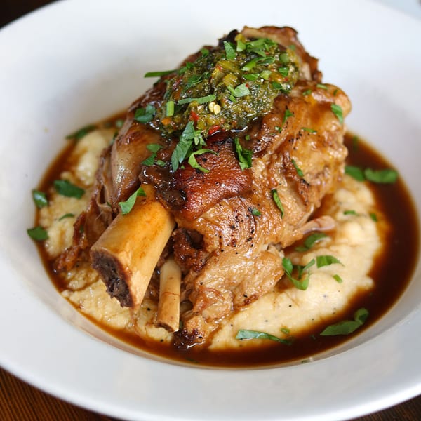 Braised pork shank with a green relish, served over polenta in a white bowl.