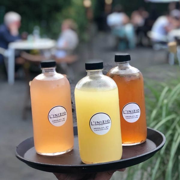 Three L'inizio branded bottles of orange, yellow, and amber drinks on a tray at an outdoor restaurant.