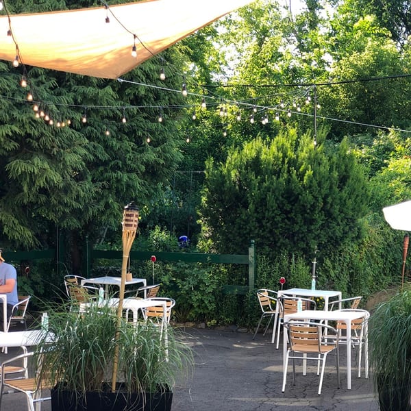 Outdoor patio with white tables, chairs, string lights, and lush green foliage under a canopy.