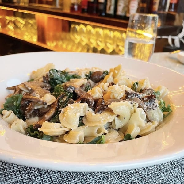 Pasta with mushrooms, kale, and parmesan cheese in a white bowl, with a blurry bar in the background.