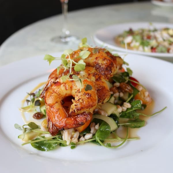 Grilled shrimp on a bed of greens, cucumber, and grains, drizzled with sauce.
