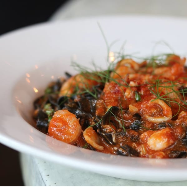 Close-up of black pasta with red sauce, shrimp, squid, and green garnish in a white bowl.