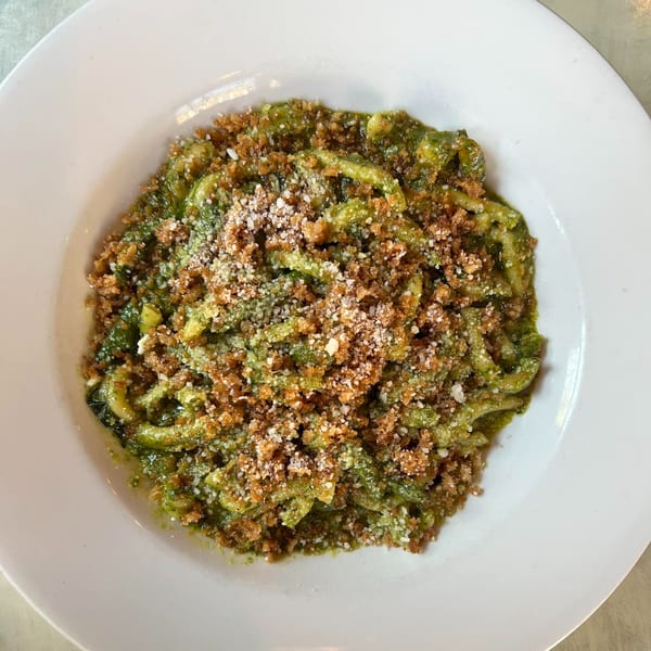Green pasta with pesto sauce, topped with breadcrumbs and grated cheese in a white bowl.