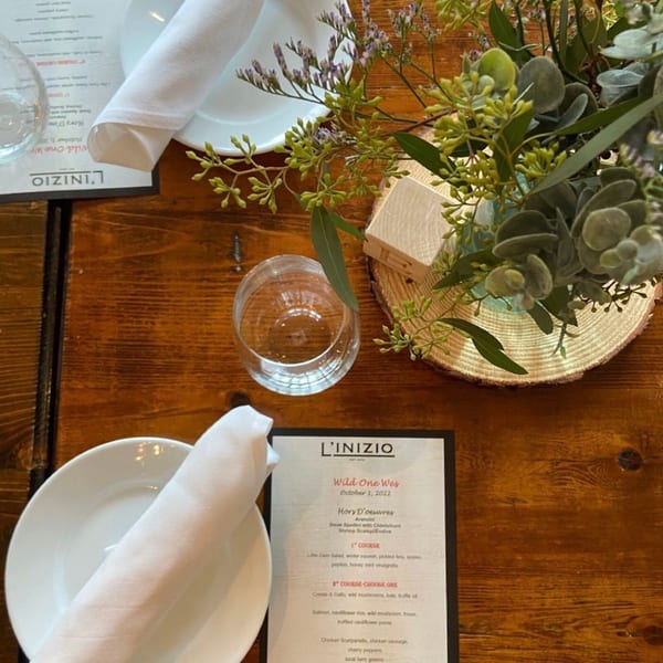 A set table at L'inizio with a menu, white plate, napkin, wine glass, and floral centerpiece.