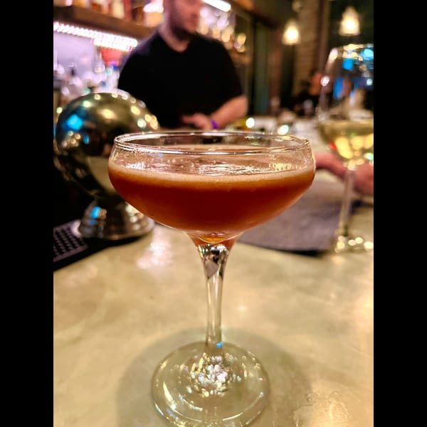 A red cocktail in a coupe glass on a marble bar, with a blurred bartender in the background.