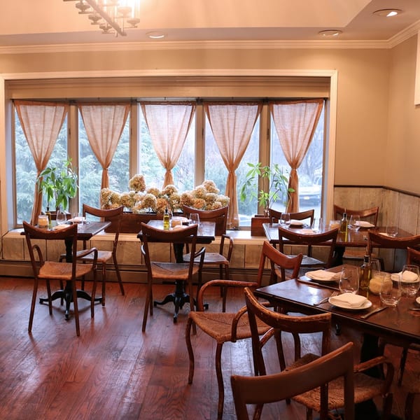 Cozy restaurant dining room with wooden tables, chairs, and large windows with sheer curtains.