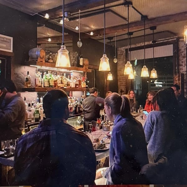 Patrons seated at a dimly lit bar with a well-stocked liquor shelf and hanging pendant lights.
