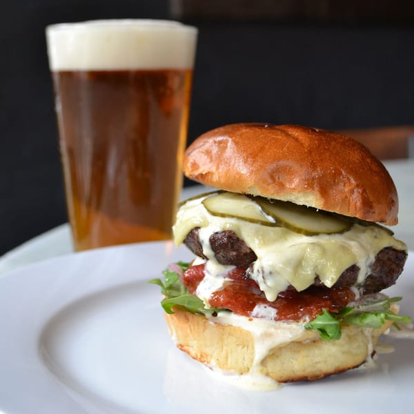 Gourmet burger with melted cheese, pickles, and sauce on a white plate, with a glass of beer.