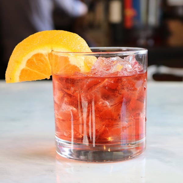 A vibrant red cocktail with ice and an orange slice garnish sits on a white marble surface.