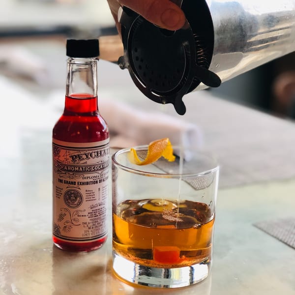 A bartender strains a cocktail into a glass with an orange peel and cherry, next to a bottle of Peychaud's Bitters.