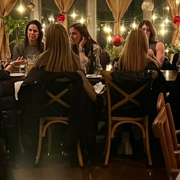 Four women dining at a restaurant table, illuminated by festive string lights and ornaments.