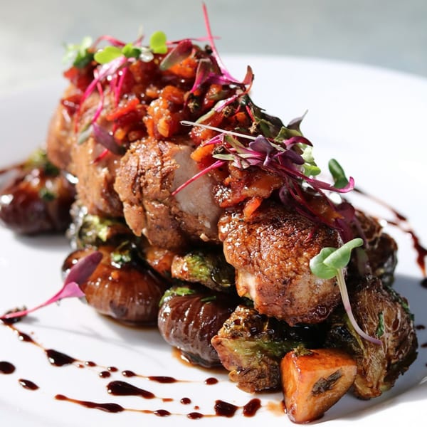 Seared pork tenderloin with roasted Brussels sprouts, pearl onions, and microgreens on a white plate.