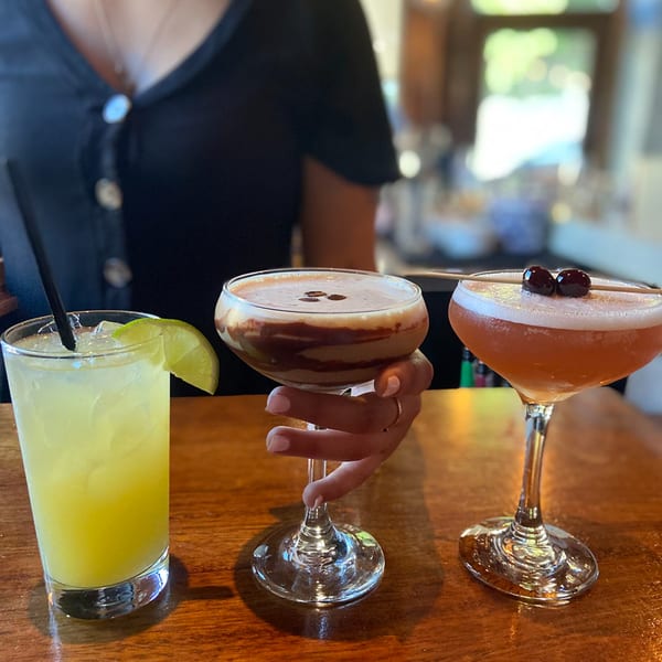 Three cocktails on a wooden bar, including a yellow drink, a chocolate martini, and a cherry-garnished drink.