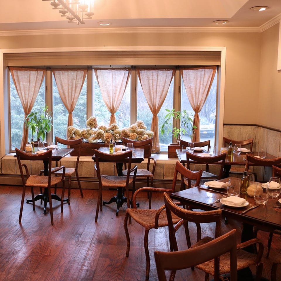 Cozy restaurant dining room with wooden tables and chairs, set against large windows with sheer curtains.