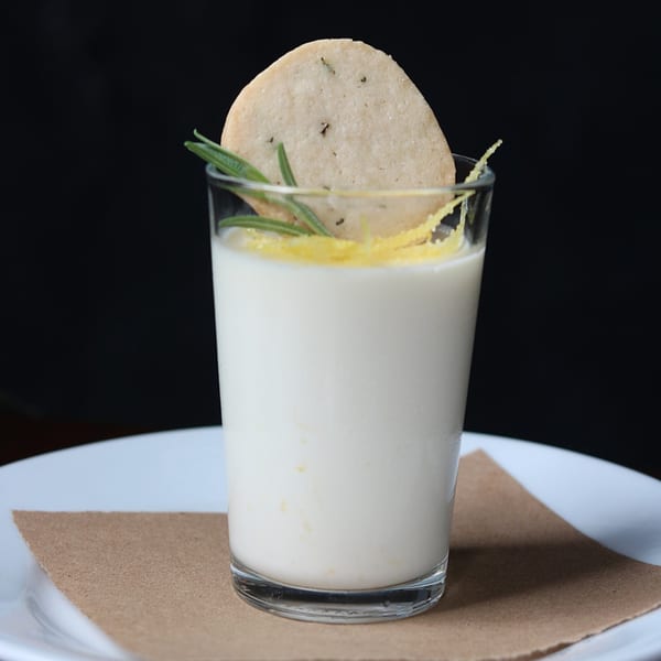 White dessert in a glass, garnished with a cookie, rosemary, and lemon zest.