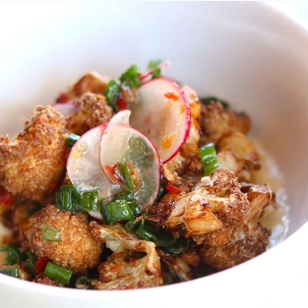 Close-up of crispy fried cauliflower florets garnished with sliced radishes and chopped green onions in a white bowl.