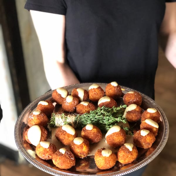 A server holds a silver tray of golden-brown arancini balls topped with white sauce and fresh herbs.