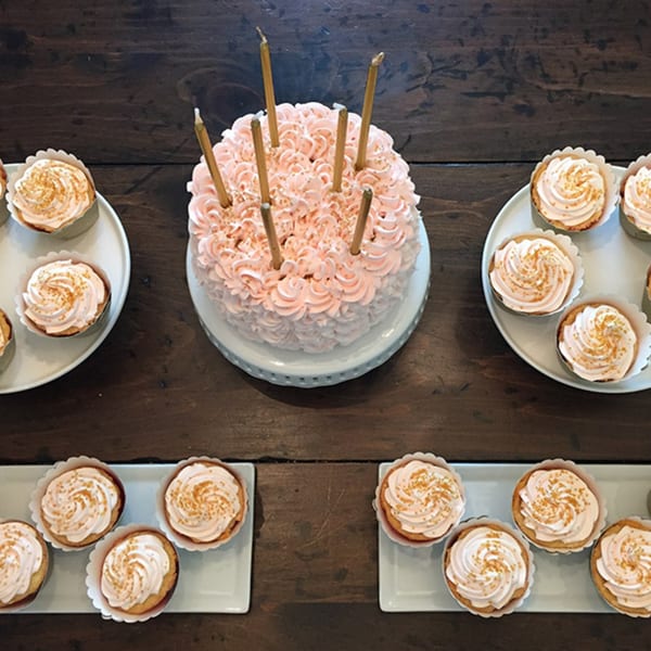Pink frosted cake with candles surrounded by cupcakes on a dark wooden table.
