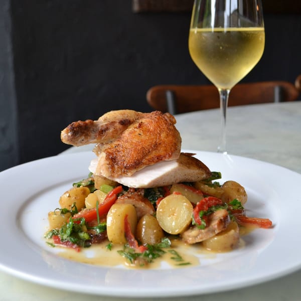 Roasted chicken with crispy skin, sliced and served over potatoes and red peppers, with a glass of white wine.