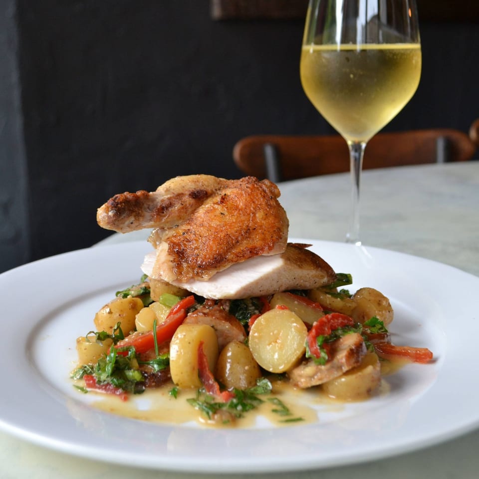 Roasted chicken with crispy skin, sliced and served over potatoes and red peppers, with a glass of white wine.